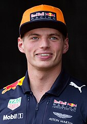 A man in his late teens twenties wearing wearing a blue sporting polo with sponsors logos. He is wearing an orange cap and is smiling