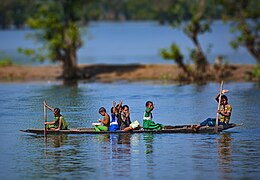 Sur une sorte de pirogue très basse, naviguant sur une rivière bleue, 7 enfants disent bonjour au photographe.