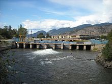 Photo d'un barrage prise depuis un lac, qui semble faire une vingtaine de mètres de large, un portail métallique est visible sous le barrage, une rambarde et un grillage bleu se trouvent au dessus. Derrière le barrage de bâtiments et des montagnes sont visibles.