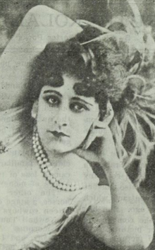 A white woman with dark curled hair and dark eyes, wearing a plummed headpiece and strands of pearls; one arm bent over her head, and her head resting on the other hand