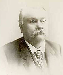 A balding, mustachioed man in his late sixties. He is wearing a black coat, white shirt, and striped tie