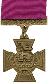 A medal in the shape of a cross hanging from a crimson red ribbon.