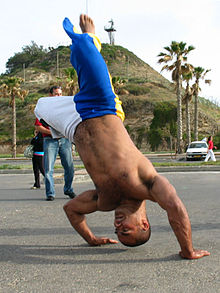 Un homme torse nu effectue un mouvement de rotation, la tête vers le sol, et se soutient par la force des bras.