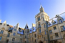 Stone buildings showing multiple windows, chimneys, towers and decorative features. Sunlight strikes the uppermost reaches, the rest is in shadow