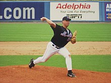 A man in a black baseball jersey and cap and gray baseball pants throws a baseball with his right hand.