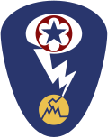 Badge d'épaule ovale avec un fond bleu foncé. Au sommet, se trouve une étoile bleue entourée d'un cercle rouge, qui correspond à l'écusson des Army Service Forces. Ce dernier est entouré d'un ovale blanc représentant un nuage en champignon. Un éclair blanc sort du nuage et frappe un cercle jaune fissuré représentant un atome.