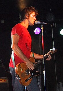 A man is shown in three-quarter side profile. He is singing into a microphone on it stand while playing a guitar. He wears a red t-shirt and jeans. Behind him are stage lights and an amplified speaker.