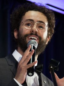 Youssef holding a microphone and smiling while wearing a tuxedo