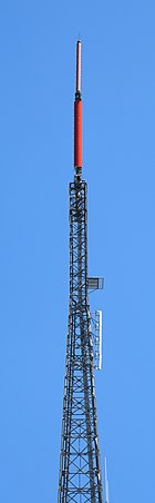 A gray lattice tower, set against a blue sky. A small pink cylindrical antenna and a larger red cylindrical antenna top the structure.