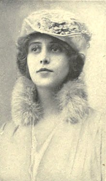 Young white woman with dark hair, wearing a light-colored hat, fur collar, and dress.
