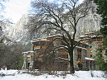A multi-story building with a wood and stone exterior is in the midground, a tree is in the foreground and high cliffs in the background. Snow is on the ground.