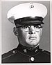 Head of a heavyset young white man with wire-frame glasses wearing a white peaked cap with black visor and a dark military jacket.