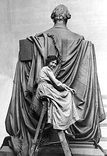 Sculptor Elsie Ward perched atop a ladder in a chamois work dress behind the sculpture which is in process