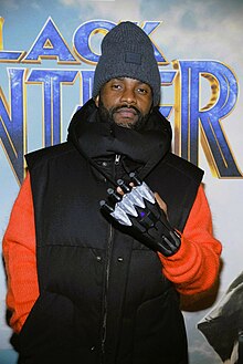 Fally Ipupa wearing a black vest over an orange sweater, a grey tuque, and a black plastic glove, looking directly at camera