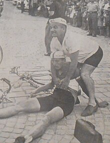 black and white image of Franc Škerlj at approximately 21 years old. He is sitting on the floor next to his bicycle and wearing cycling gear. There is a man standing over him. He looks to have fallen off his bike during a race in front of a crowd