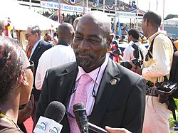 A black-skinned man with a bald head and black goatee beard. He is wearing a light blue shirt with the top buttons undone, showing a white top underneath.