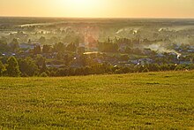 Photographie depuis une colline (le mont Piket) montrant des maisons du village de Strotski, avec de la fumée de cheminée. Le paysage est vert, avec des arbres entre les maisons. La colline est une prairie.