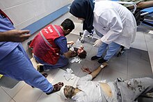 A bloodied child on the floor of a hospital being treated by a nurse. A man with his head wrapped in a bandage wearing blood-stained clothes lays on the floor nearby.