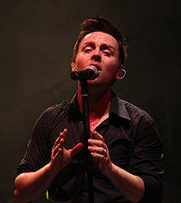 A dark-haired young man in a dark shirt singing into a microphone