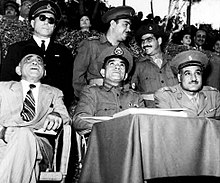 Three men seated and observing an event. The first man from the left is wearing a suit and fez, the second man is wearing a military uniform, and the third man is wearing military uniform with a cap. Behind them are three men standing, all dressed in military uniform. In the background is ab audience seated in bleachers