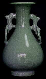 A tall, thin, blue-green vase with two handles, each with a small upper opening and a larger lower opening.