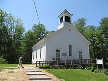 A small white church building, with trees in the background. A man walks towards thr church.