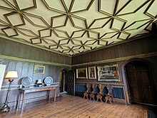 A Tudor room, with dark wood-paneled walls and intricate ceiling woodwork, containing a number of paintings and pieces of furniture