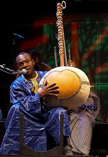 Toumani Diabaté wearing a dark blue African outfit, holding a kora, sitting on a chair onstage and speaking into a microphone