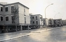 Black and white photograph showing a boulevard with trees and streetlamps lined with white houses on stilts.