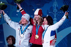 Three smiling women stand side by side with one arm over each other's back. Each holds high a flower bouquet and wears a medal around the neck. On the left, a long-haired blond wears a shiny light-gray jacket and winter cap with a badge bearing the Norwegian flag and Olympic rings. On the center, another long-haired blond wears a red jacket and red-and-white winter cap. On the right, a long-haired brunette wears a white jacket with blue and red stripes on the back.