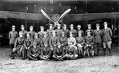Twenty-five uniformed men standing or seated in front of a biplane with a four-bladed propeller
