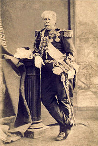 Full-length photographic portrait depicting an older man with moustache resting his right arm on a pedestal which holds a feathered bicorne hat, and wearing a heavily decorated military dress uniform with his left hand resting on the sword at his side