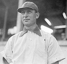 A man in a dark baseball cap facing 3/4 towards the camera. A baseball stadium grandstand appears to be in the background.