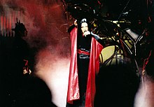 Madonna with short brunette hair wearing a black-and-red kimono and singing into a microphone.