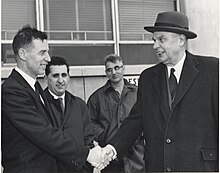Diefenbaker, wearing a coat over his suit, shakes hands with a smiling man. Two other men are in the background.