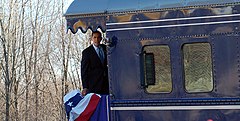 Obama is standing aboard the back platform of a train and looking to the side of the train. There is a red, white, and blue banner hanging over the rear railing.