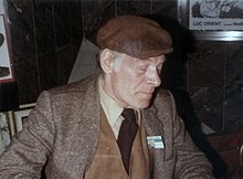 Photographie d'un homme assis portant un costume, un béret, se tenant les yeux fermés.