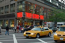 A street view of a store front prominently features a yellow taxi in front of the store. The walls contain clear windows with the phrase "Tower Records – Video" in bright orange lights.
