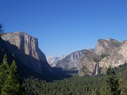 Mountain valley with sheer cliff on its left side, and a waterfall cascading into its right, with a clear blue sky above and many green trees below