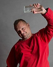 Jurgen Teller wearing a red sweatshirt with his left arm raised holding an acrylic award