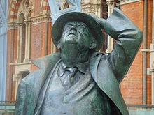 A bronze statue of an old man, wearing a suit and tie and an open long coat, looking up and holding a hat on his head with his left hand; behind, part of a brick building with Gothic arched windows