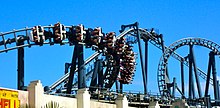 A train loaded with passengers plummets through a banked turn on Lethal Weapon – The Ride; the coaster's lift hill and rollover inversion are in the background.