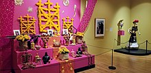 Photo of museum exhibits. At left, a brightly colored three-tiered altar featuring reminders of lost loved ones like food, sculptures, and photographs. At right, a skeleton wears a dark dress and bright flower hat.