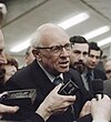 An elderly man with receding white hair and large spectacles, Andrei Sakharov, is being interviewed. A tape recorder is held in front of his mouth by a hand from the bottom of the photograph. Sakharov is wearing a suit with a blue and brown striped tie.