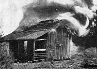 A black and white photograph of a crude wooden structure that could be a small shed, animal house, or hunting cabin with smoke pouring from it and flames visible in the door