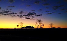 Small, pointed mountain on the horizon, foregrounded by scattered bare trees, silhouetted against the sky at sunset