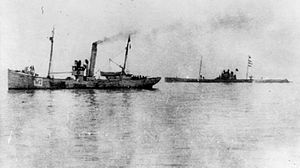 A submarine on the surface in the middle distance on the right. Nearer to the camera, on the left, is a small steam vessel.
