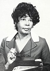 Photograph of an African-American woman dressed in a suit with wide lapels, holding her glasses in front of her in her right hand and a book in her left hand.