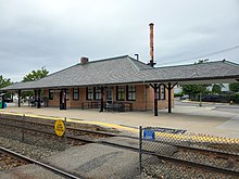 A one-story yellow brick station building next to a rail line. The roofline connects to a canopy which extends both directions along the platform.