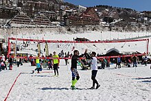 Two competitors stand between a net above the snowy playing field with a crowd a cabin and a snowy mountain in the backround